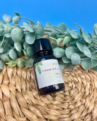 Thymes Frasier Fir Refresher Oil in amber glass bottle with signature evergreen label displayed on a solid blue background.