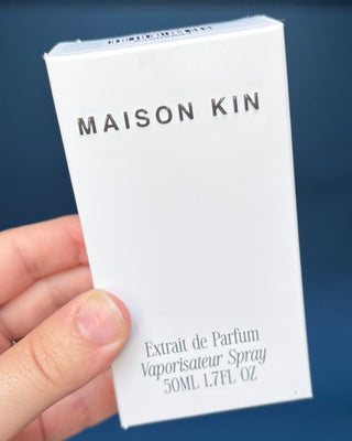Maison Kin perfume packaging held by a hand against a blue background