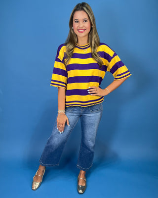 A woman modeling a yellow and purple striped short-sleeve knit top with blue jeans and metallic flats, standing against a solid blue studio backdrop.