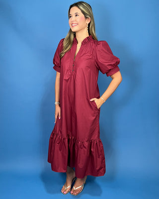 A woman modeling a short-sleeve midi dress in a deep burgundy color, standing against a solid blue studio backdrop.