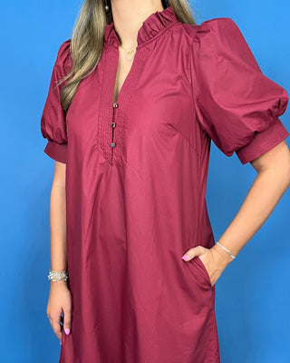 A woman wearing a dusty rose puff-sleeve tunic dress with a ruffled neckline and button placket, standing against a solid blue background.