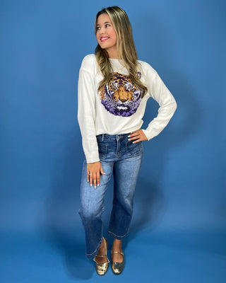 A woman modeling a cream-colored crewneck sweater with a large, detailed tiger face graphic on the front in purple, gold,