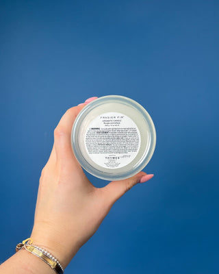 Thymes Frasier Fir Tidings 13 oz. candle in a decorative gift box displayed against a solid blue background. 
