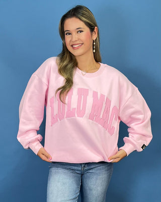 A woman wearing a light pink crewneck sweatshirt with 'LULU MAC' printed in arched pink letters across the chest, paired with blue jean