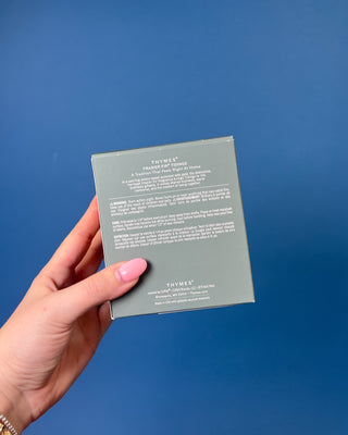 Thymes Frasier Fir Tidings 13 oz. candle in a decorative gift box displayed against a solid blue background. 