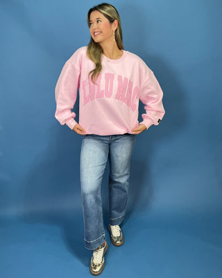 A woman modeling a light pink crewneck sweatshirt with 'LULU MAC printed in darker pink across the chest, paired with blue jeans and metallic sneaker