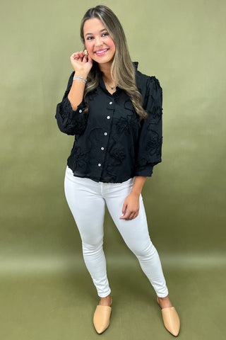 Woman wearing a black blouse with floral details and white pants on a green background