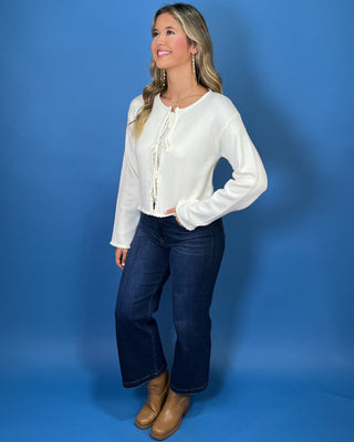 Ivory knit cardigan top with long sleeves, double front tie closures, and delicate ruffle trim along the edges.