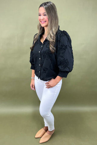Woman wearing a black blouse with floral details and white pants on a green background