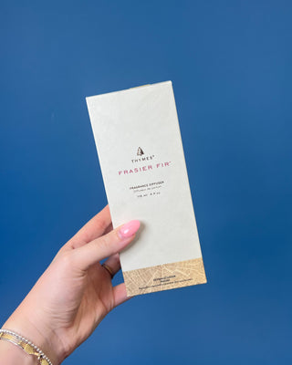 Thymes Frasier Fir Petite Gold Diffuser with reeds displayed on a solid blue background. 