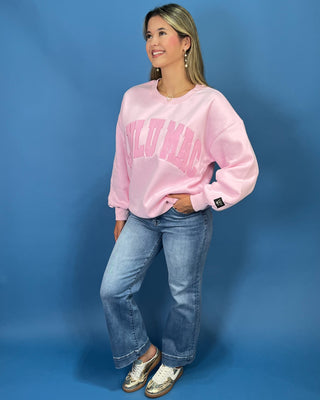 A woman modeling a light pink crewneck sweatshirt with 'LULU MAC printed in darker pink across the chest, paired with blue jeans and metallic sneaker