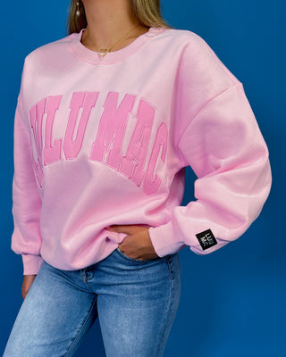 A woman modeling a light pink crewneck sweatshirt with 'LULU MAC printed in darker pink across the chest, paired with blue jeans and metallic sneaker