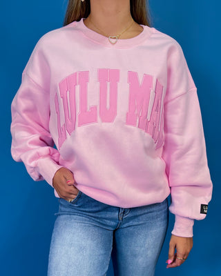 A woman modeling a light pink crewneck sweatshirt with 'LULU MAC printed in darker pink across the chest, paired with blue jeans and metallic sneaker