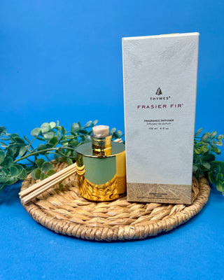Thymes Frasier Fir Petite Gold Diffuser with reeds displayed on a solid blue background. 