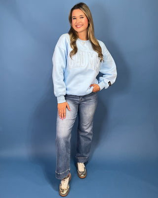 A woman modeling a light blue sweatshirt with 'LULU MAC' printed across the chest, paired with blue jeans and gold sneaker