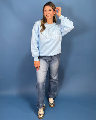 A woman modeling a light blue sweatshirt with 'LULU MAC' printed across the chest, paired with blue jeans and gold sneaker