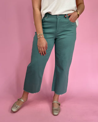 Person wearing teal pants and a white top on a pink background