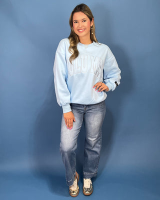 A woman modeling a light blue sweatshirt with 'LULU MAC' printed across the chest, paired with blue jeans and gold sneaker