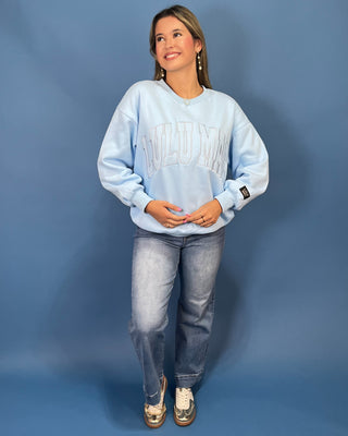 A woman modeling a light blue sweatshirt with 'LULU MAC' printed across the chest, paired with blue jeans and gold sneaker