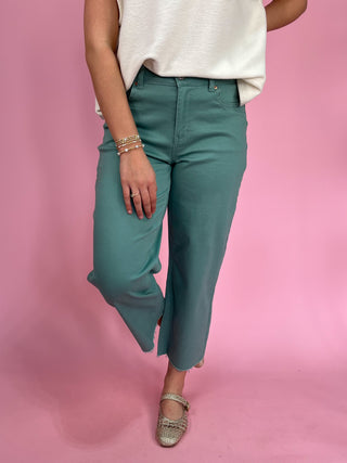Person wearing teal pants and a white top on a pink background