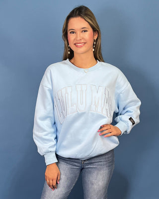 A woman modeling a light blue sweatshirt with 'LULU MAC' printed across the chest, paired with blue jeans and gold sneaker