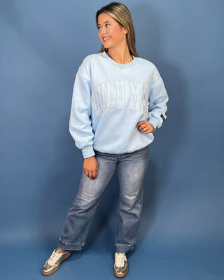 A woman modeling a light blue sweatshirt with 'LULU MAC' printed across the chest, paired with blue jeans and gold sneaker