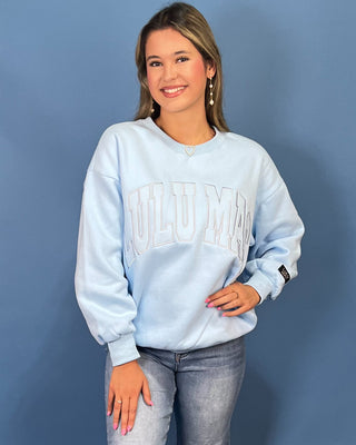 A woman modeling a light blue sweatshirt with 'LULU MAC' printed across the chest, paired with blue jeans and gold sneaker