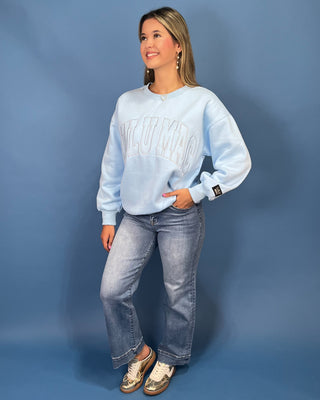 A woman modeling a light blue sweatshirt with 'LULU MAC' printed across the chest, paired with blue jeans and gold sneaker