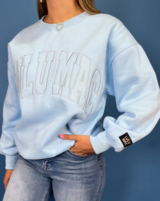 A woman modeling a light blue sweatshirt with 'LULU MAC' printed across the chest, paired with blue jeans and gold sneaker