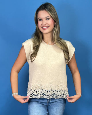 A woman modeling a cream-colored sleeveless top with a lace hem, paired with blue jeans and metallic gold flat