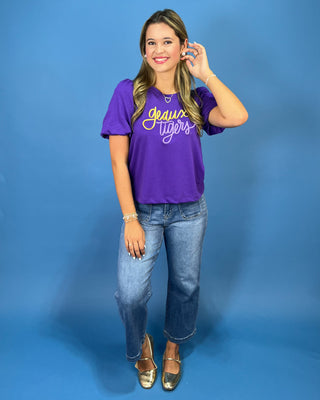 A woman modeling a purple short-sleeve graphic t-shirt with 'Gravitas Tigers' written in yellow script across the chest, paired with blue jeans and metallic gold flats, set against a solid blue studio backdrop.