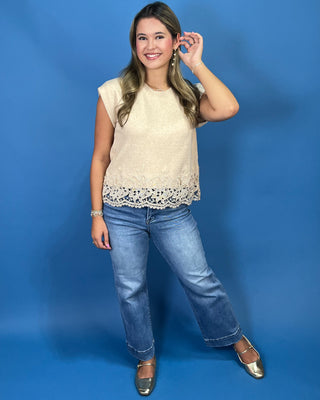 A woman modeling a cream-colored sleeveless top with a lace hem, paired with blue jeans and metallic flats