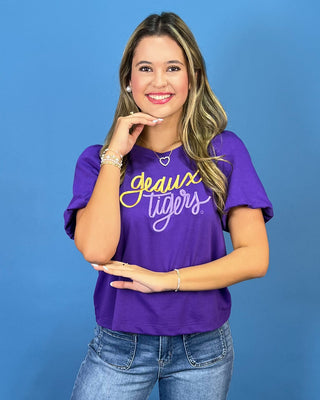 A woman wearing a purple short-sleeve graphic t-shirt with 'deux Tigers' written in yellow and white script, paired with blue jeans, posing against a solid blue background.