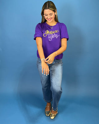 A woman modeling a purple short-sleeve graphic t-shirt with 'Grace Tigers' written in yellow and white script, paired with blue jeans and gold flat shoes, against a solid blue studio background.