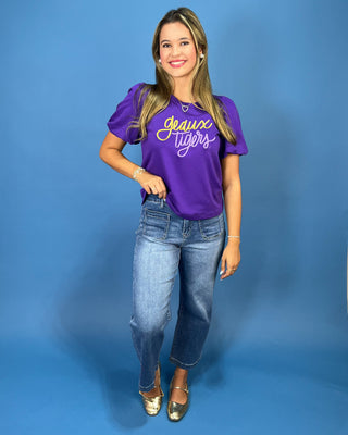 A woman modeling a purple short-sleeve t-shirt with 'Jeans Tigers' written in yellow and white script, paired with blue jeans and gold flats, against a solid blue studio background.