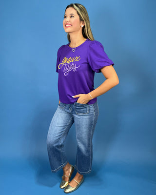 A woman modeling a purple short-sleeve t-shirt with 'Juice Jocks' written in yellow and white script across the chest, paired with blue jeans and gold flats, against a solid blue studio background.