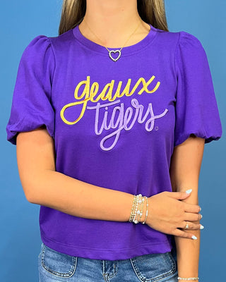 A woman wearing a purple short-sleeve t-shirt with 'geaux tigers' printed in yellow and white script across the chest, paired with blue jeans against a solid blue background.