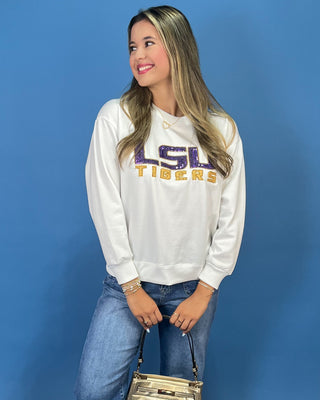 A woman wearing a white LSU Tigers sweatshirt with purple and gold lettering, paired with blue jeans, standing against a solid blue background.