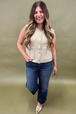 Woman wearing a beige sleeveless cardigan and blue jeans against a green background