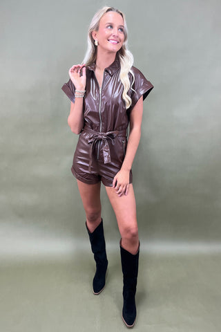 Woman wearing a shiny brown romper with black knee-high boots on a plain background