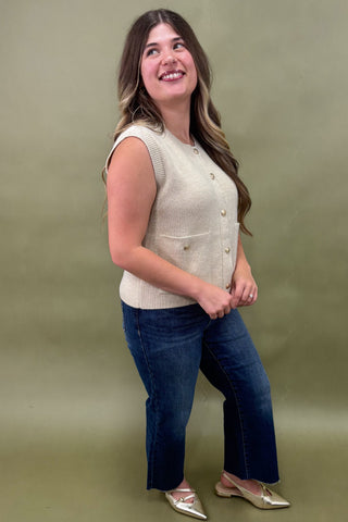 Woman wearing a beige sleeveless vest and blue jeans against a green background