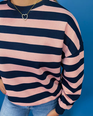 A close-up of a person wearing a pink and navy blue horizontally striped crewneck sweatshirt, paired with light blue jeans and a gold heart necklace