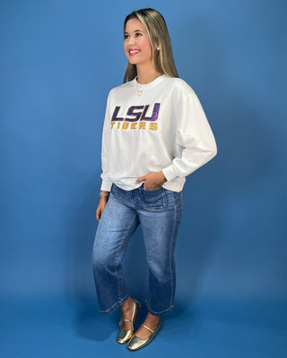 A woman modeling a white LSU Tigers sweatshirt with purple and gold lettering, paired with blue jeans and gold flats, standing against a solid blue studio backdrop.