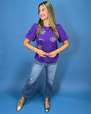 A woman modeling a purple short-sleeve t-shirt with gold and white graphic prints, paired with blue jeans and metallic flats, standing against a solid blue studio backdrop.