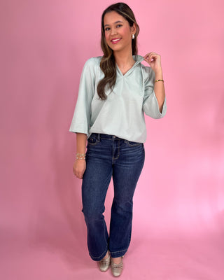 Woman wearing a light green blouse and blue jeans against a pink background