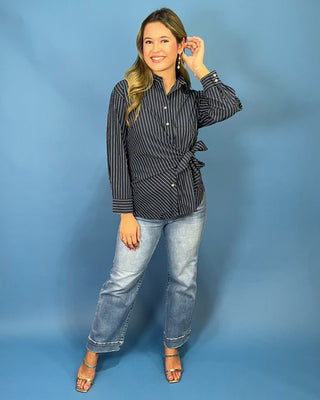 A woman modeling a black and white vertically striped wrap-style button-up shirt with a side tie detail, paired with light-wash blue jeans