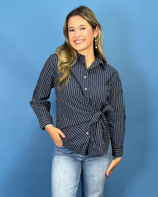 A woman modeling a navy blue and white vertically striped button-down shirt with a wrap-style front and side tie detail, paired with light-wash jean