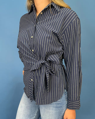 A woman wearing a navy blue long-sleeve button-up shirt with thin white vertical pinstripes and a self-tie waist, paired with light-wash blue jean