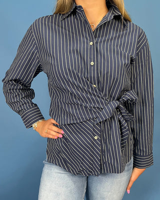 A woman wearing a navy blue and white pinstriped wrap-style button-up shirt with a tie waist, paired with light-wash jean