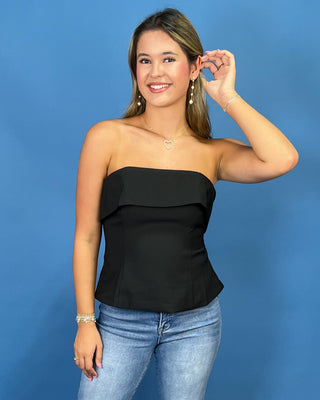 A woman modeling a black strapless peplum top with ruffle detailing, paired with light wash blue jeans